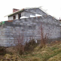 Budowa domu z betonu komórkowego w stanie surowym otwartym z widoczną ścianą boczną, fragmentem dachu i kominem z cegły, na pierwszym planie sucha trawa i krzewy bez liści.